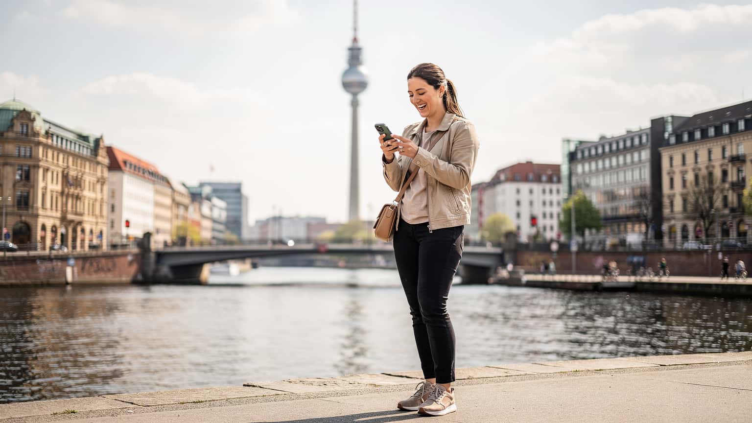 Eine junge Frau, gekleidet für einen Städtetrip, lacht, während sie am Ufer der Spree in Berlin-Mitte steht und ihr Smartphone mit der digitalen Karte einer Stadtansicht in den Händen hält, mit dem Fernsehturm im Hintergrund.