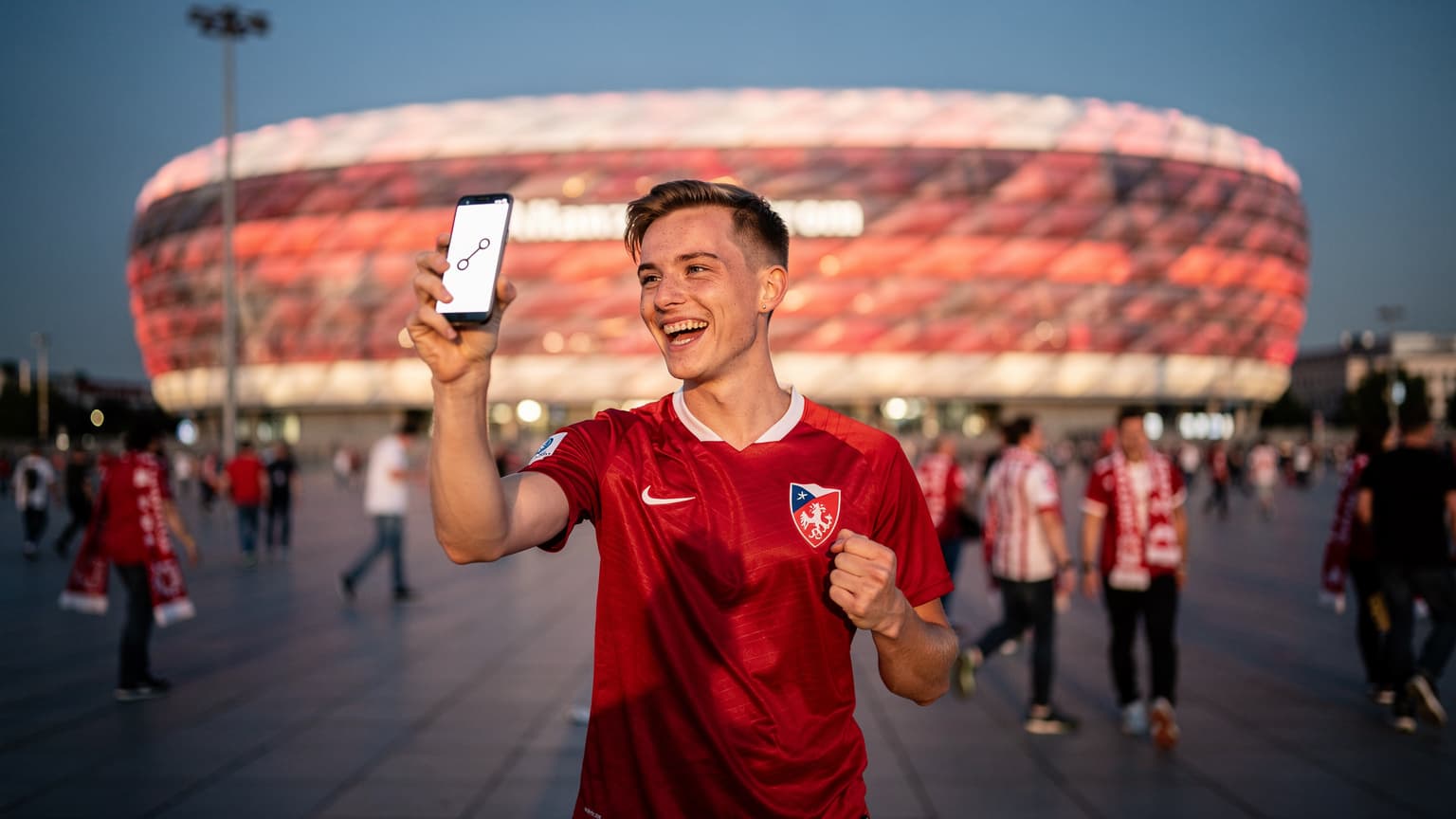 Mladý český fanoušek s dresem české reprezentace se usmívá a drží telefon s aktivní eSIM kartou před osvětleným stadionem v Mnichově.