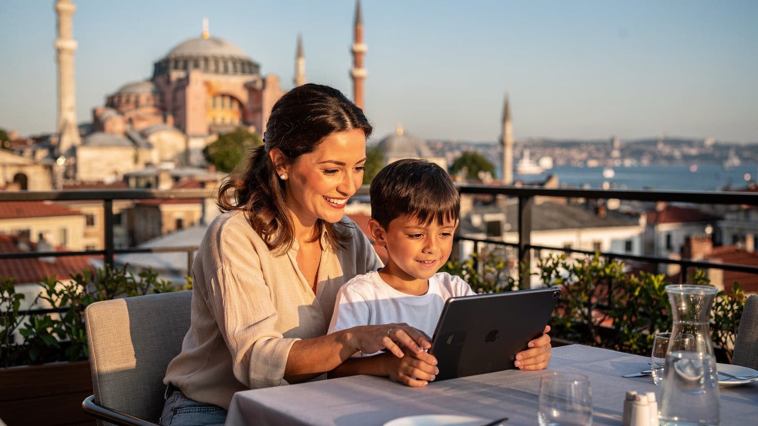 O mamă zâmbește în timp ce își ajută fiul să-și seteze tableta pe terasa unui restaurant de pe acoperiș din Istanbul, cu o vedere panoramică spre Hagia Sofia și Bosfor.