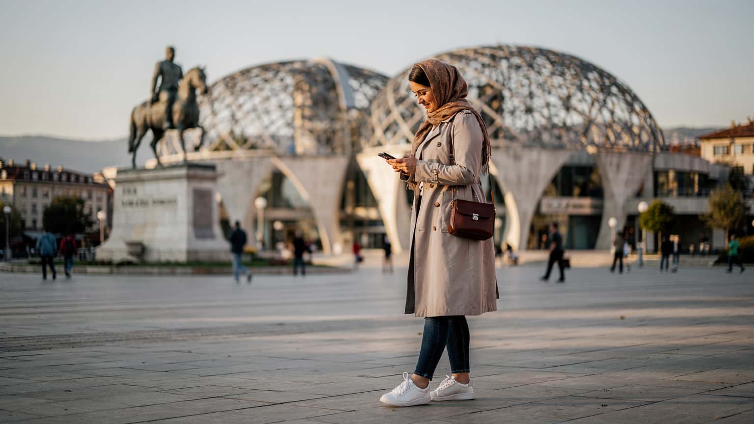 Priştine'deki İskenderbey Meydanı'nda bir kadın, elindeki telefonla haritaya bakarken gülümsüyor, arka planda Ulusal Kütüphane'nin kendine özgü mimarisi seçiliyor.