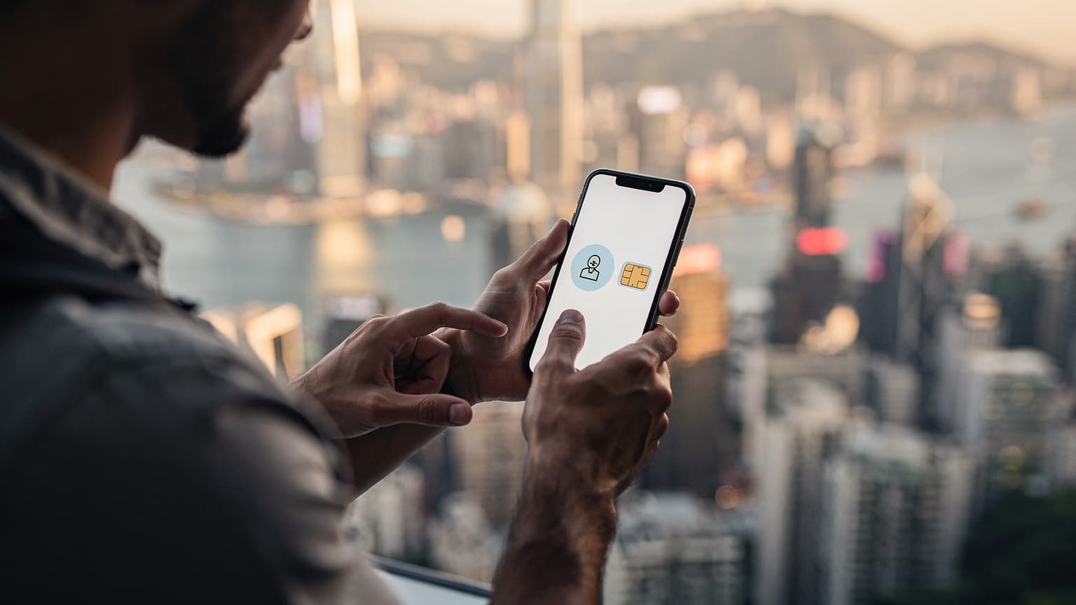 A traveler holds a smartphone against a blurred Hong Kong skyline at sunset, subtly comparing digital and physical SIM options.