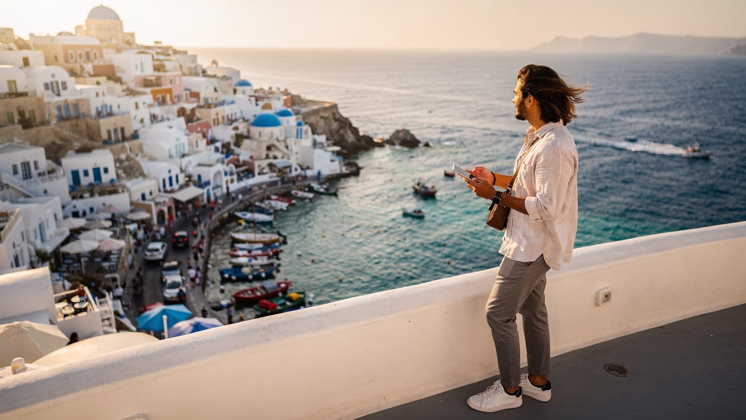 A hero image showing a person on a sun-drenched Greek island balcony with white architecture and the Aegean Sea. The person holds a smartphone, symbolizing connectivity, with a blurred Greek city harbor in the background, representing both island and city travel.