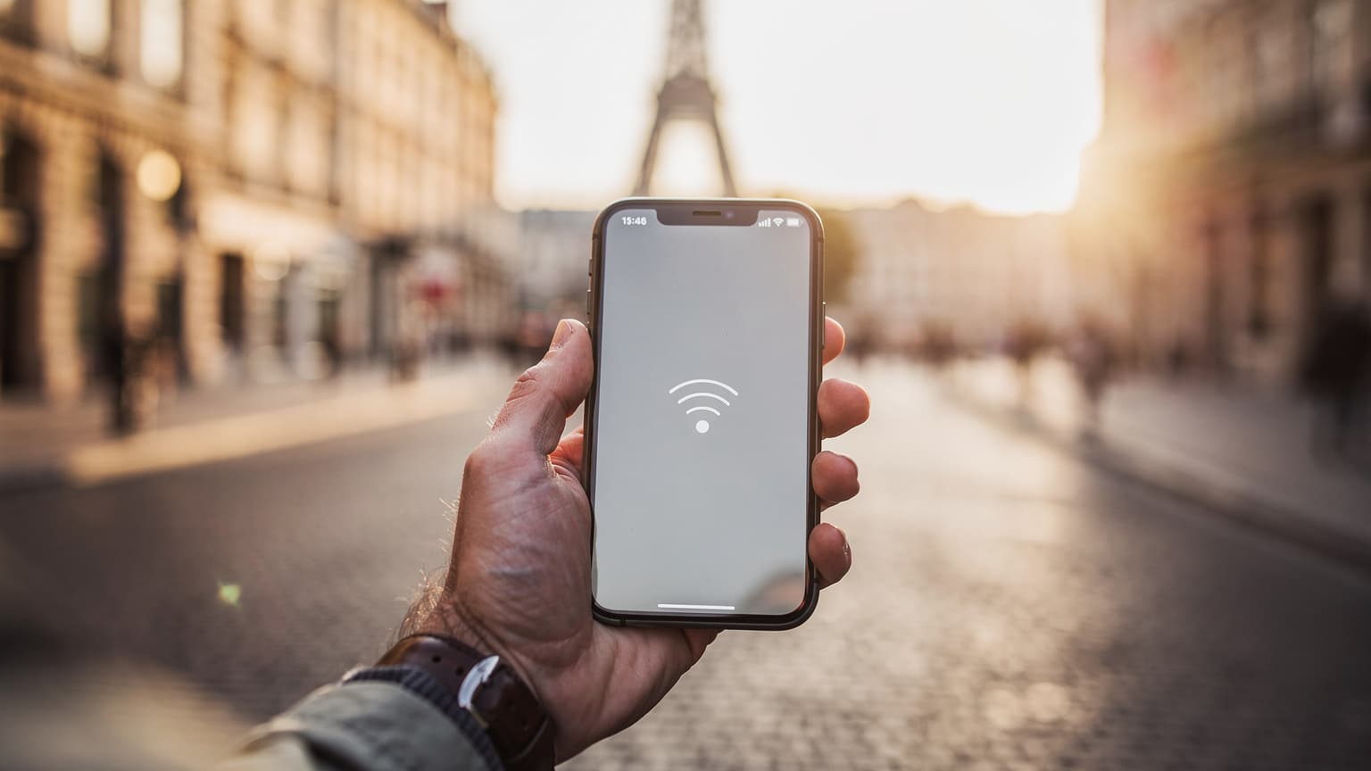 A traveler's hand holds a modern smartphone displaying a generic network signal icon, set against a blurred background of a sunny, iconic European city street, emphasizing global connectivity while traveling.