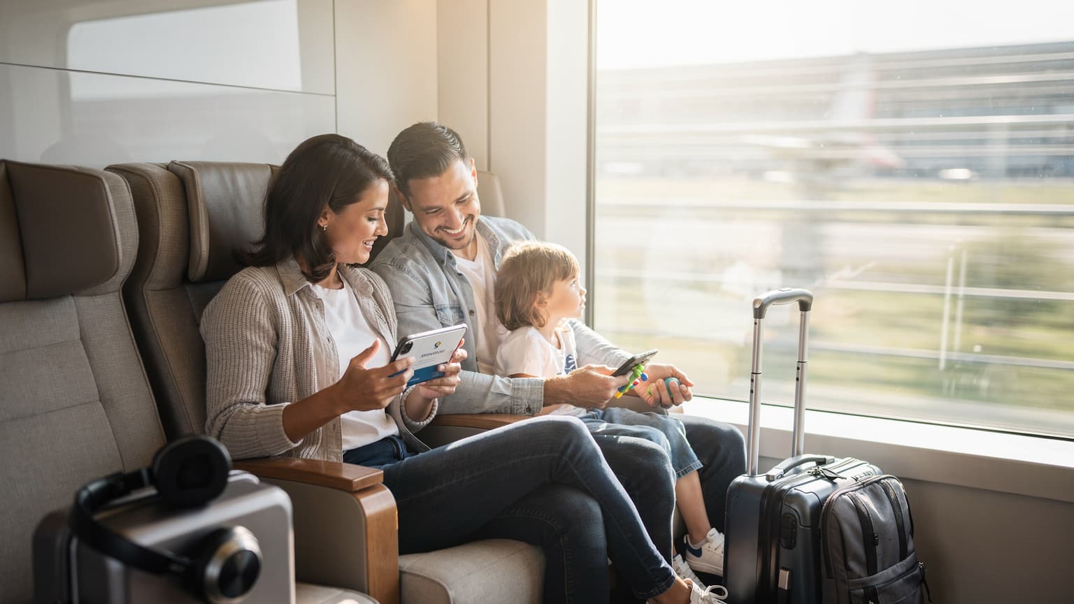 A modern family, with parents and two young children, relaxing in a bright airport lounge or train. One parent is using a smartphone, symbolizing digital travel convenience like eSIMs, while the children are contentedly engaged with their surroundings. The image conveys ease and connectivity during family travel.