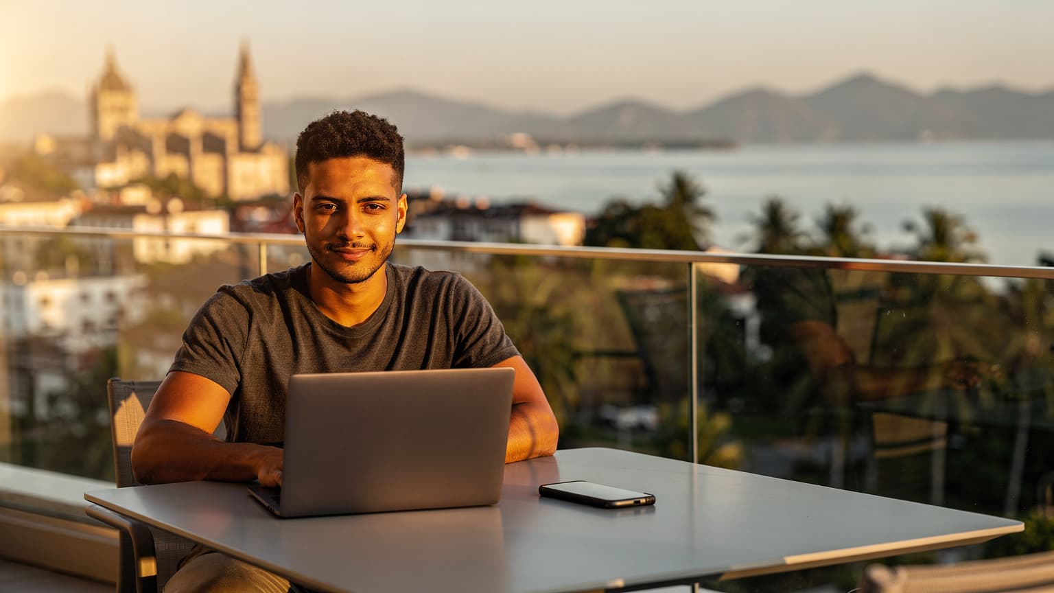 A digital nomad working on a laptop on a balcony overlooking a beautiful city or coastal landscape at sunset, symbolizing global travel and connectivity.