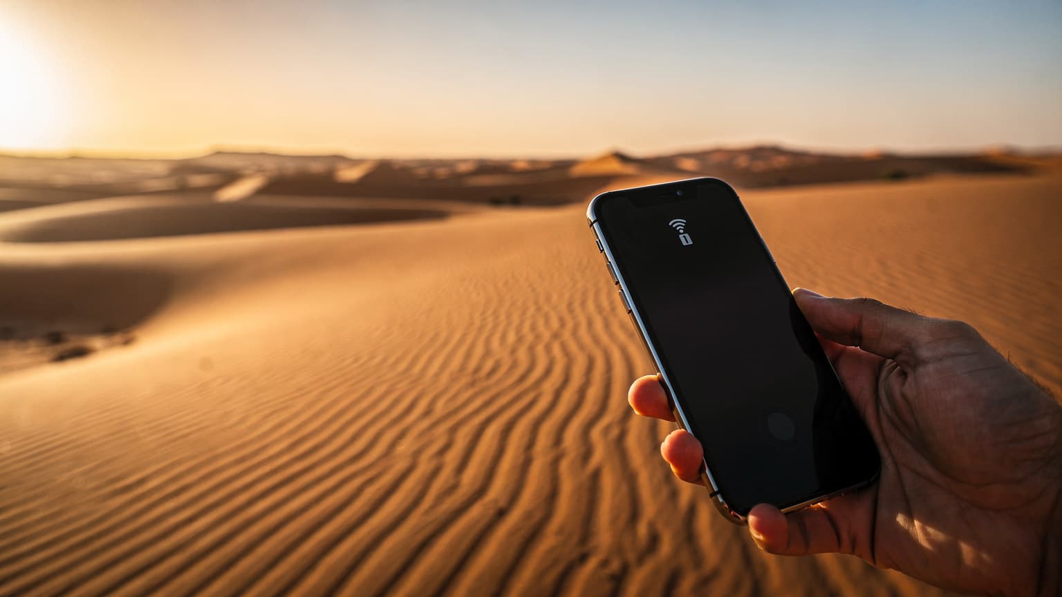 A photorealistic hero image depicting a hand holding a modern smartphone against a backdrop of vast, sunlit golden sand dunes in the Moroccan desert during golden hour, symbolizing fast and convenient internet connectivity for exploring remote areas.