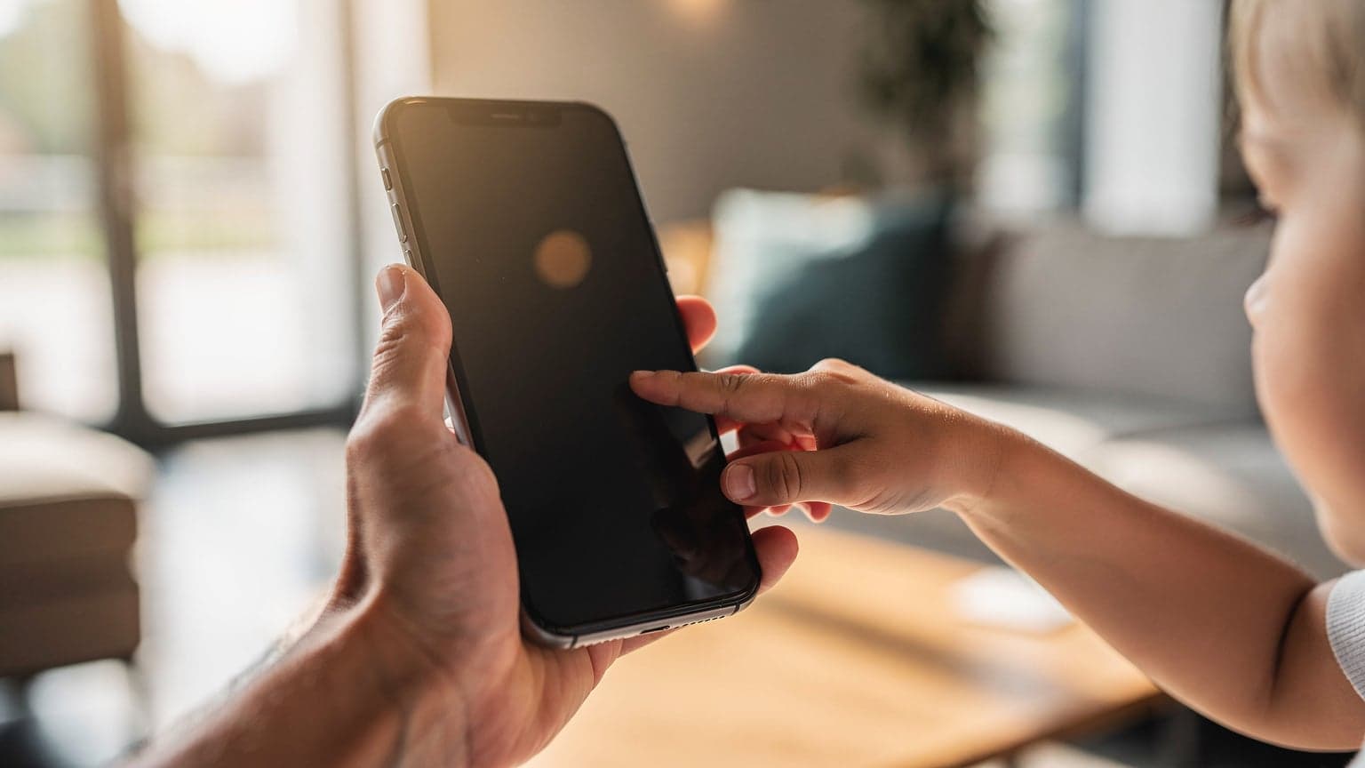 A modern smartphone held by an adult, with a child's hand touching the screen, in a bright, family-friendly setting, representing family connectivity and technology.