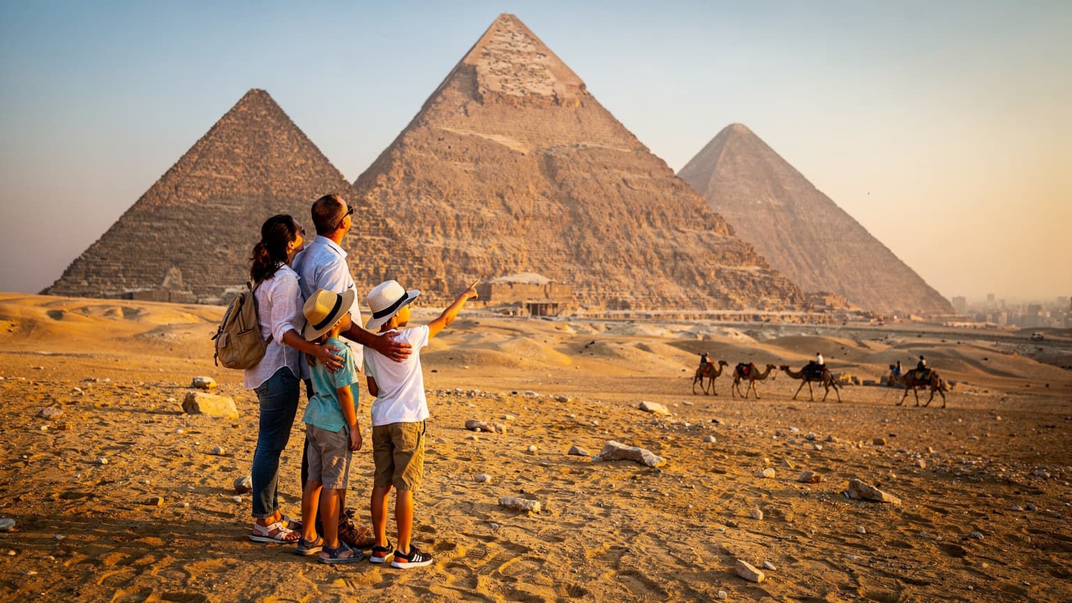 O familie cu doi copii, cu vârste între 8 și 12 ani, admirând piramidele din Giza la apus, cu un fundal de deșert auriu și o caravană mică de cămile în depărtare. Lumina caldă a soarelui cade pe fețele lor zâmbitoare.