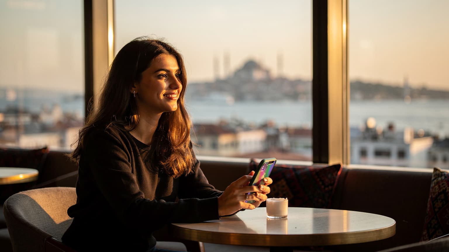 O tânără româncă zâmbește în timp ce privește spre Bosfor dintr-o cafenea modernă de pe acoperișul unei clădiri din cartierul Karaköy, Istanbul, cu telefonul său cu eSIM pregătit pentru călătorie.