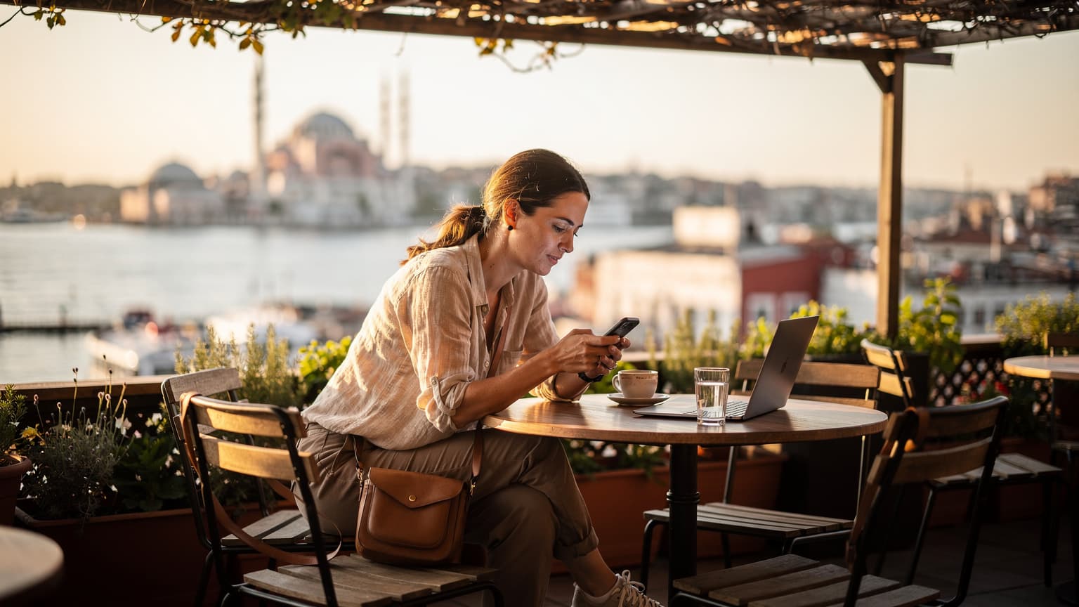 O femeie navigând pe telefonul ei în timp ce stă la o masă pe o terasă însorită cu vedere la Bosfor, în Istanbul.