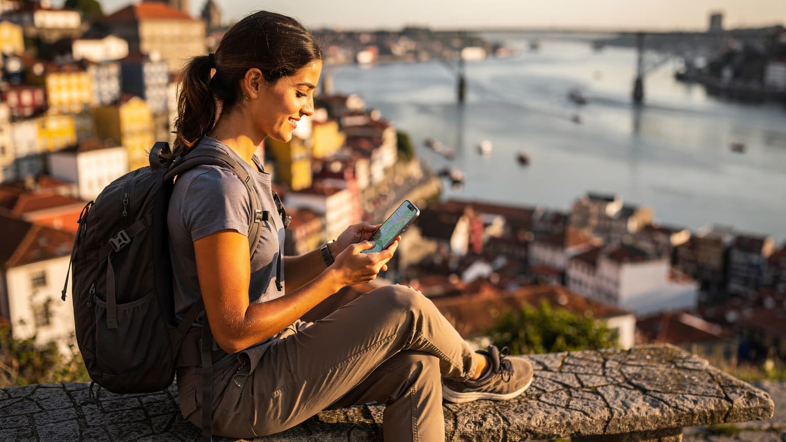 Uma jovem mochileira sorri enquanto verifica seu smartphone com um aplicativo de mapa aberto, sentada em um banco de pedra em um miradouro com vista para o Rio Douro e as casas coloridas do Porto, Portugal, ao pôr do sol.
