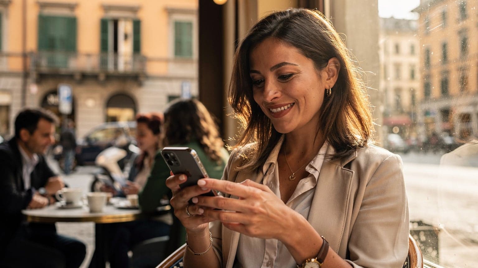En kvinde sidder ved et bord på en travl italiensk café i Milano, kigger på sin iPhone med et smil, mens hun drikker en cappuccino. Hendes bagage står diskret ved siden af hende.