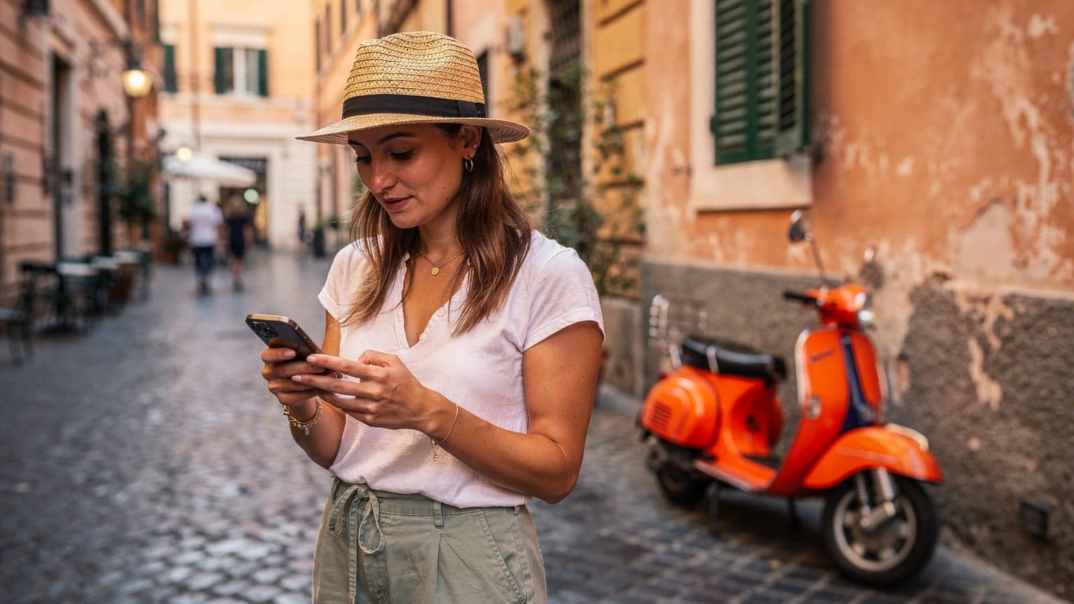 A woman holding a smartphone, looking at a digital map while standing on a cobblestone street in Rome, with a vespa parked nearby and ancient architecture in the background.