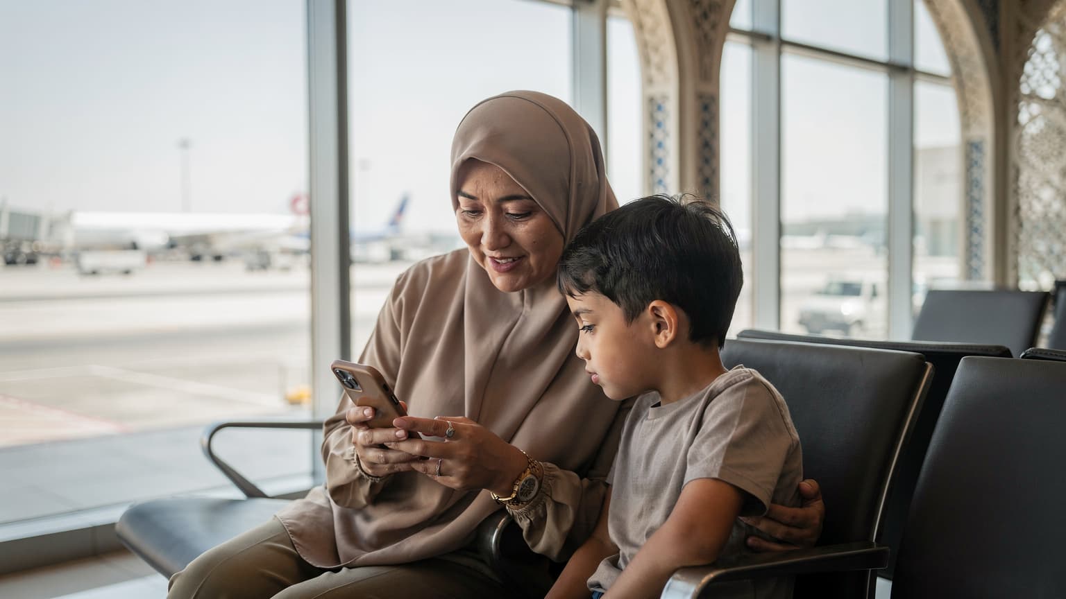 Seorang ibu dengan hijab dan anaknya melihat layar ponsel di area tunggu Bandara Internasional King Abdulaziz Jeddah, sambil memegang paspor dan tas kecil.
