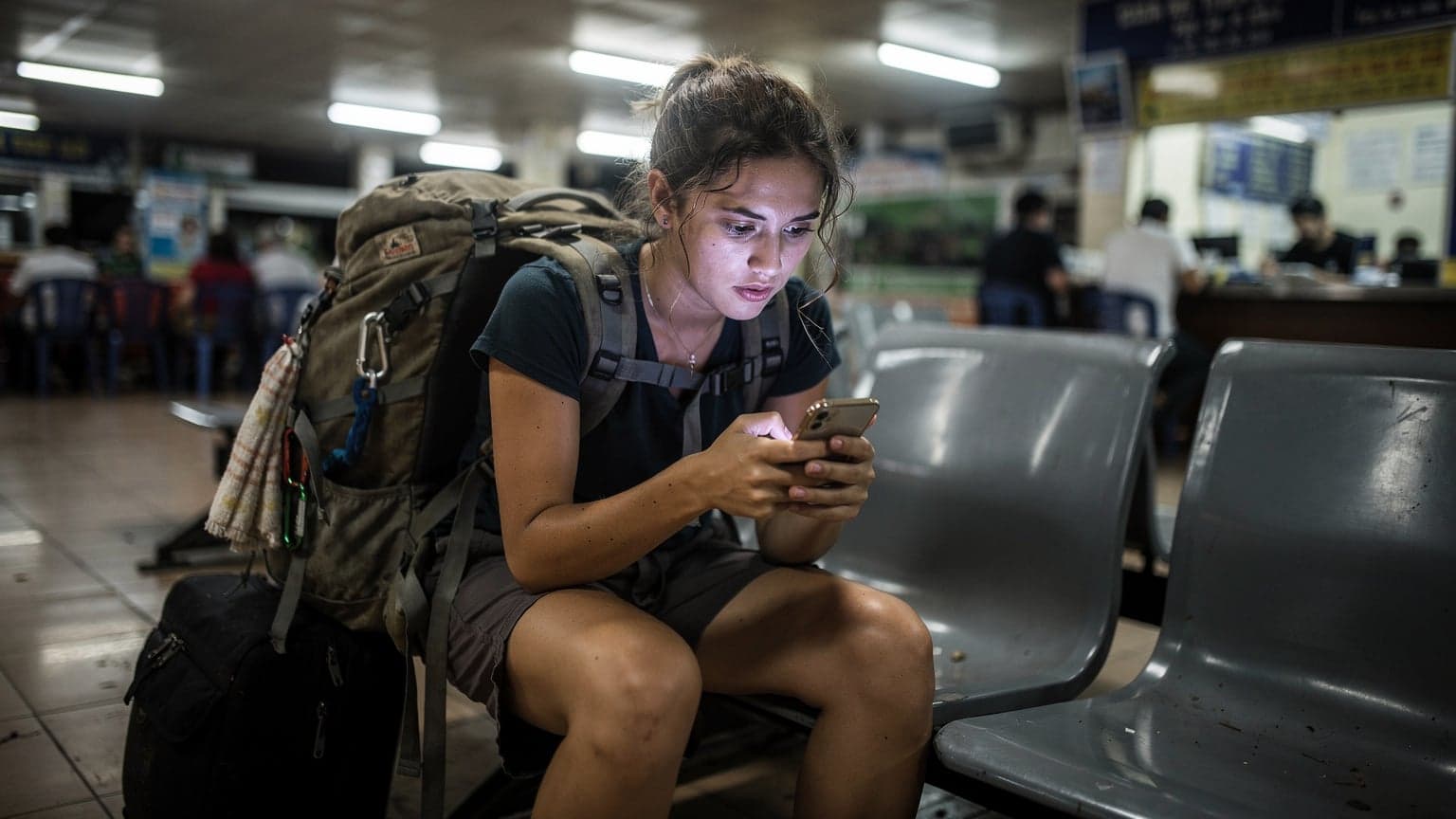 A budget backpacker reviews eSIM options on her phone while waiting for a night bus at a dimly lit bus terminal in Southeast Asia, with her large backpack at her feet.