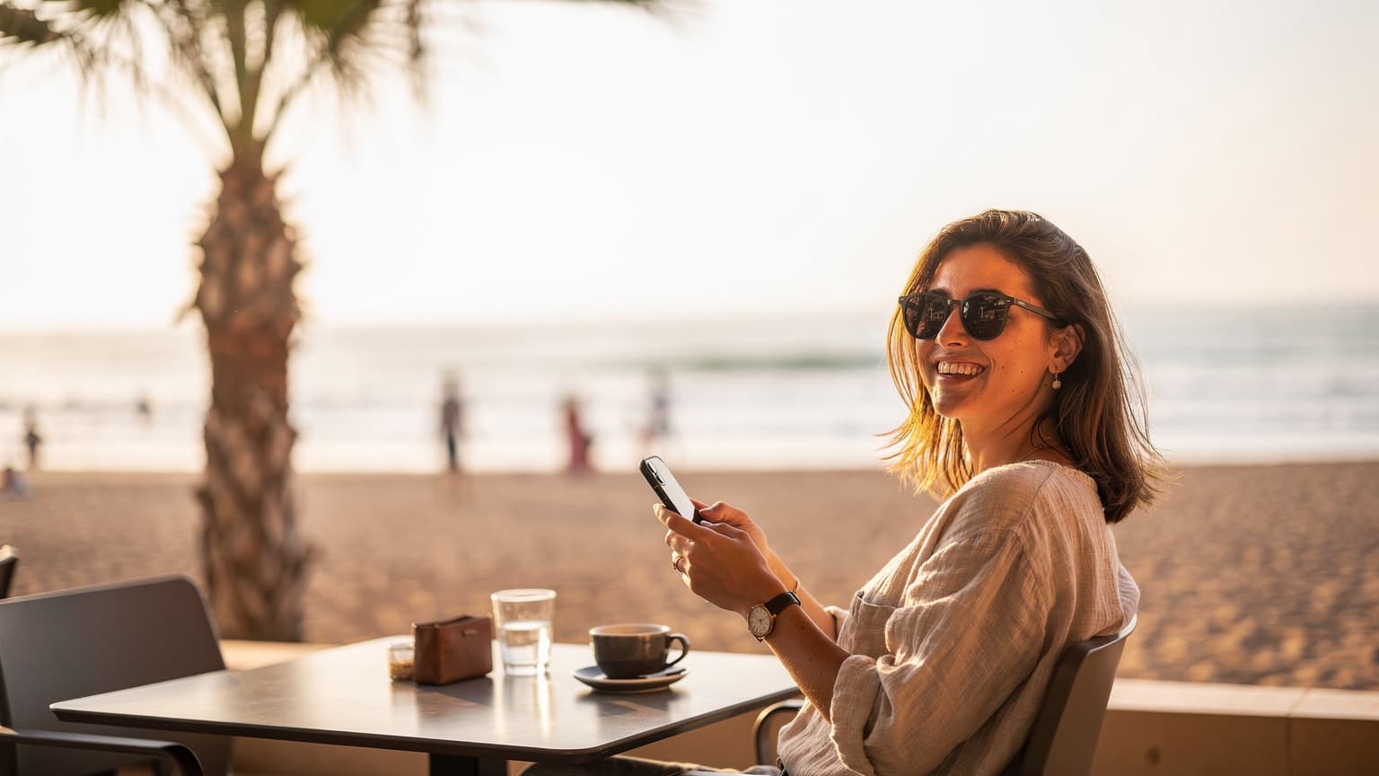 Młoda kobieta w okularach przeciwsłonecznych, trzymająca smartfon i uśmiechająca się, siedzi na tarasie kawiarni z widokiem na plażę w Agadirze w Maroku, a w tle widać palmę i ocean.