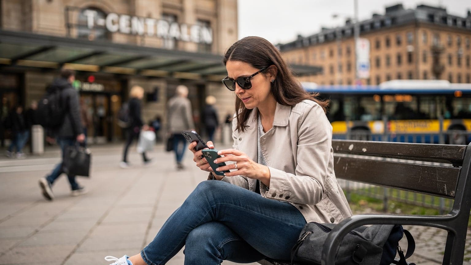 En kvinna i trettioårsåldern, med solglasögon, håller sin nya smartphone med en hand och sin gamla med den andra, sittandes på en bänk vid T-Centralen i Stockholm under rusningstid, fokuserad på att flytta över data.