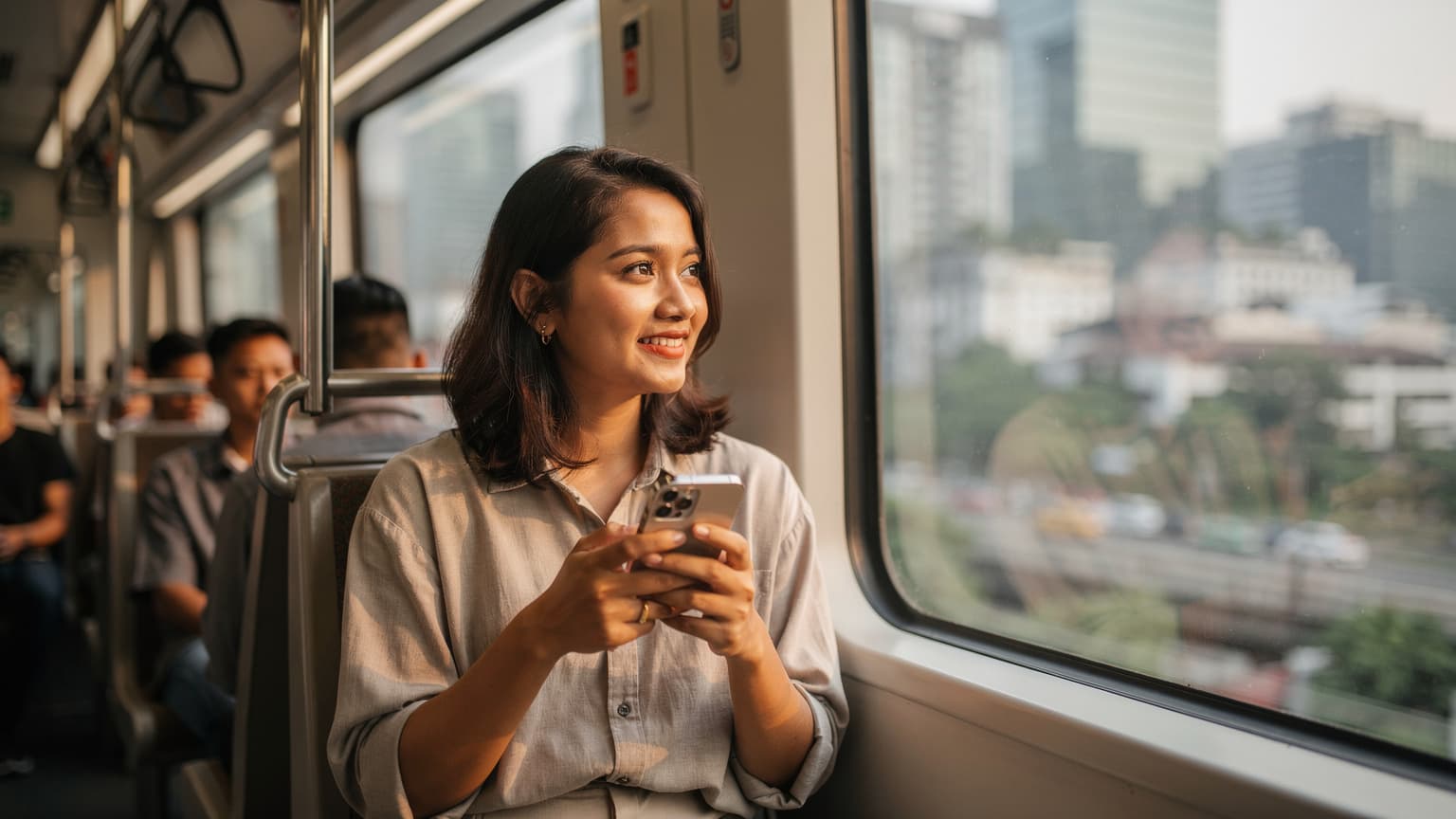 Seorang wanita tersenyum sambil memegang ponselnya di depan jendela kereta komuter yang sedang bergerak melintasi distrik perkantoran Jakarta, dengan latar belakang gedung-gedung tinggi.