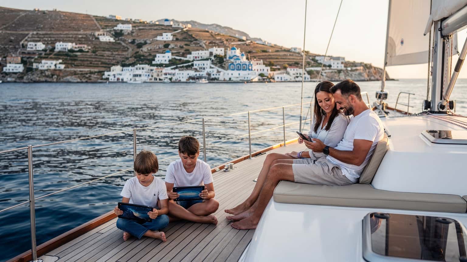 En familj med två barn, cirka 8 och 10 år gamla, sitter på en segelbåtens däck, tittar på surfplattor medan de seglar i det lugna, soliga Egeiska havet med vita kykladiska hus i bakgrunden.