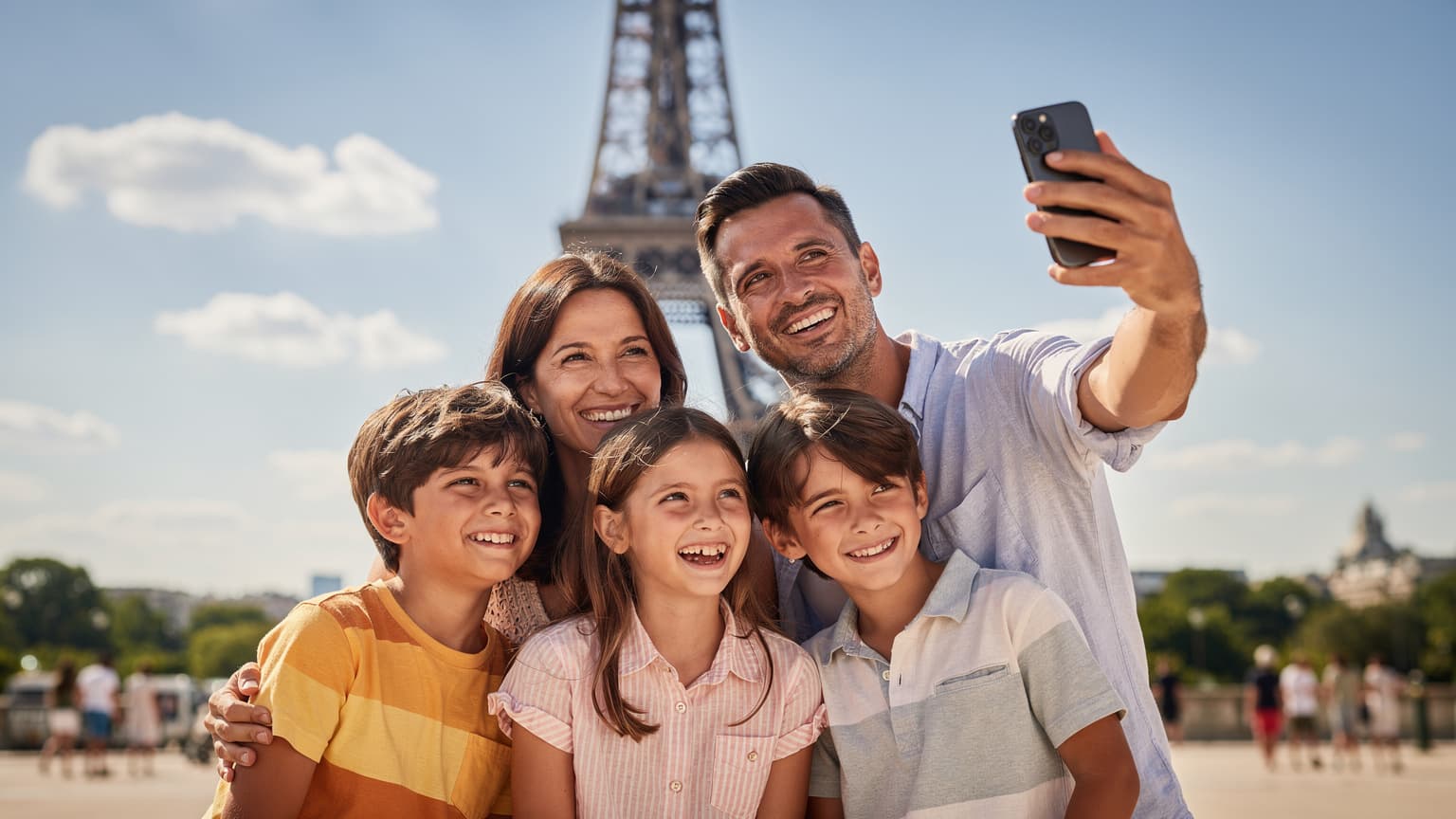 Una famiglia italiana, genitori e due bambini, sorride mentre usa gli smartphone per scattare una foto ricordo davanti alla Torre Eiffel a Parigi, con un cielo azzurro e soleggiato.