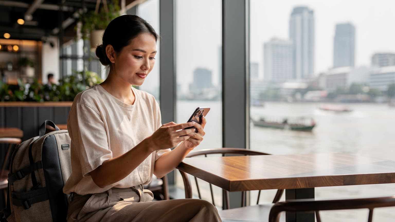一名女性在曼谷Iconsiam购物中心内的True Digital Park咖啡馆里,使用最新的iPhone 18手机连接eSIM,背景是湄南河的现代城市景观。