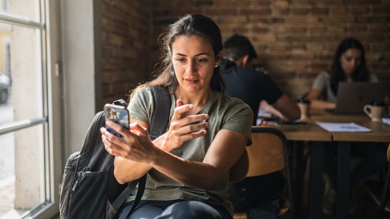 A digital nomad sits at a wooden table in a busy co-working space in Lisbon's Cais do Sodré neighborhood, looking at her iPhone with a focused expression, a half-empty coffee cup beside her.