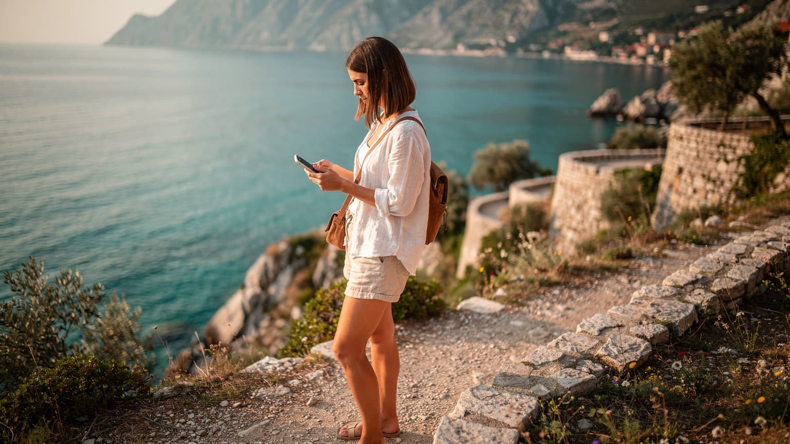 Eine junge Frau, gekleidet in leichte Reisekleidung, steht auf einem felsigen Küstenpfad mit Blick auf das klare türkisfarbene Wasser der Adria und hält ihr Smartphone in der Hand, um eine Route zu überprüfen.