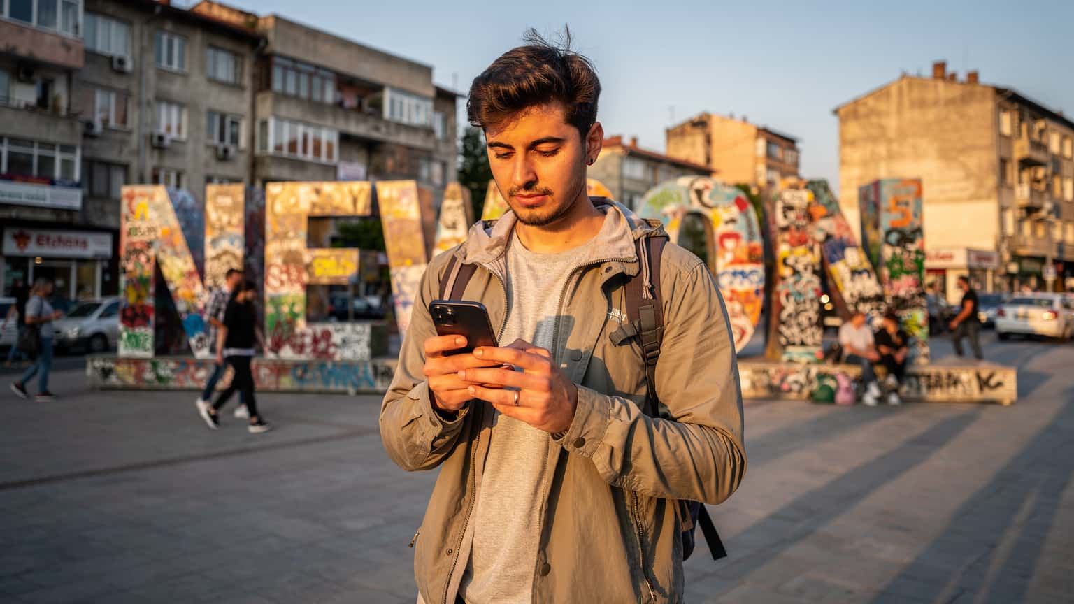 Priştine'nin 'Newborn' anıtı önünde, elinde telefonla eSIM kurulumu yapan genç bir dijital göçebe, gün batımı ışığında.