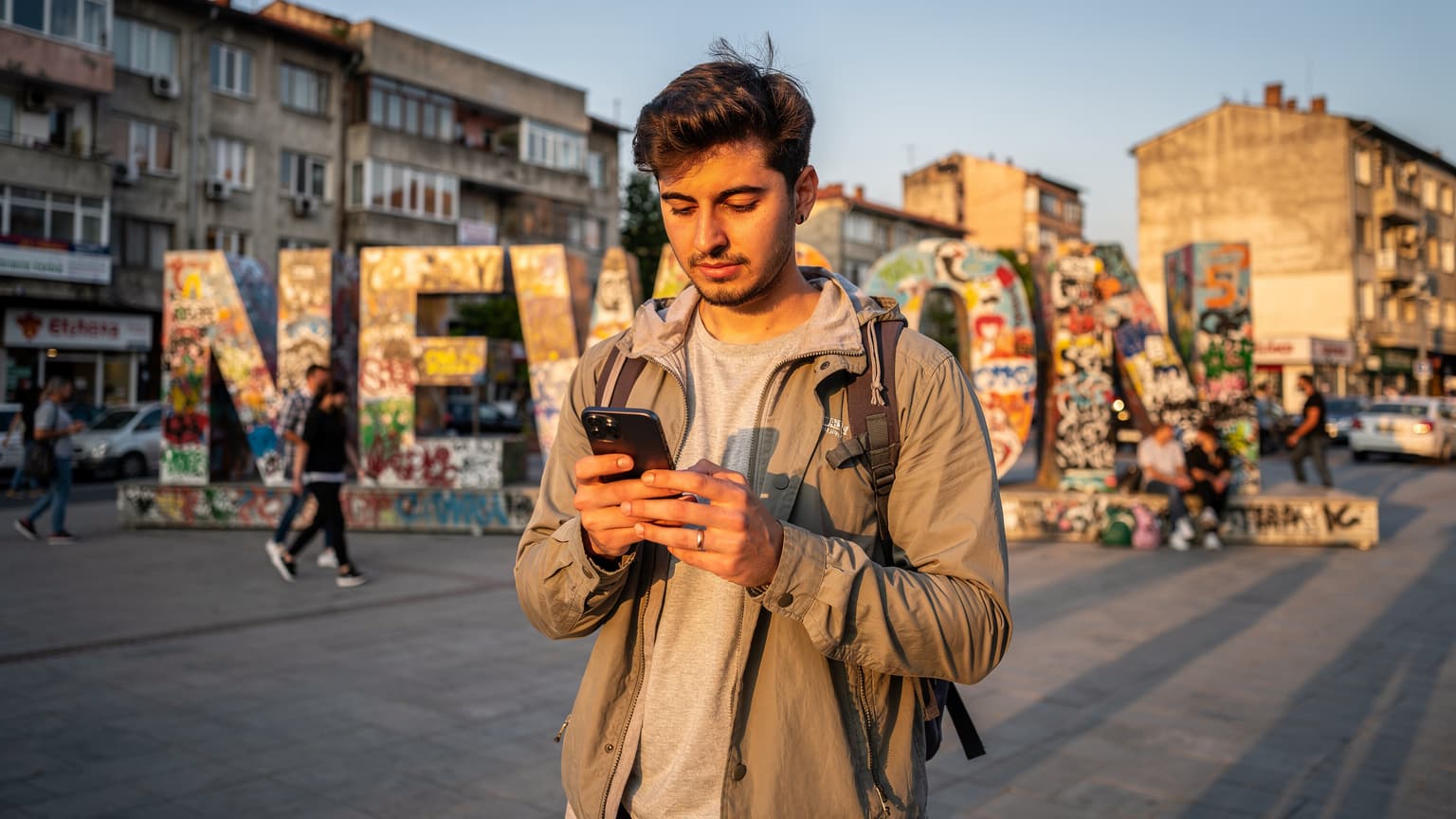 Priştine'nin 'Newborn' anıtı önünde, elinde telefonla eSIM kurulumu yapan genç bir dijital göçebe, gün batımı ışığında.