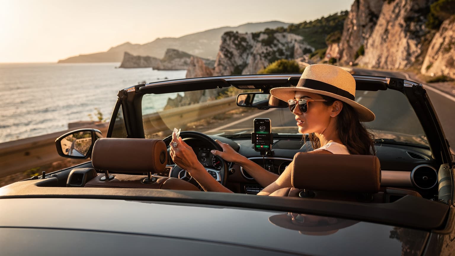 Een vrouw met een zonnebril en een breedgerande hoed navigeert op haar telefoon in een cabriolet langs de kustweg van de Côte d'Azur, met de Middellandse Zee en palmbomen op de achtergrond.