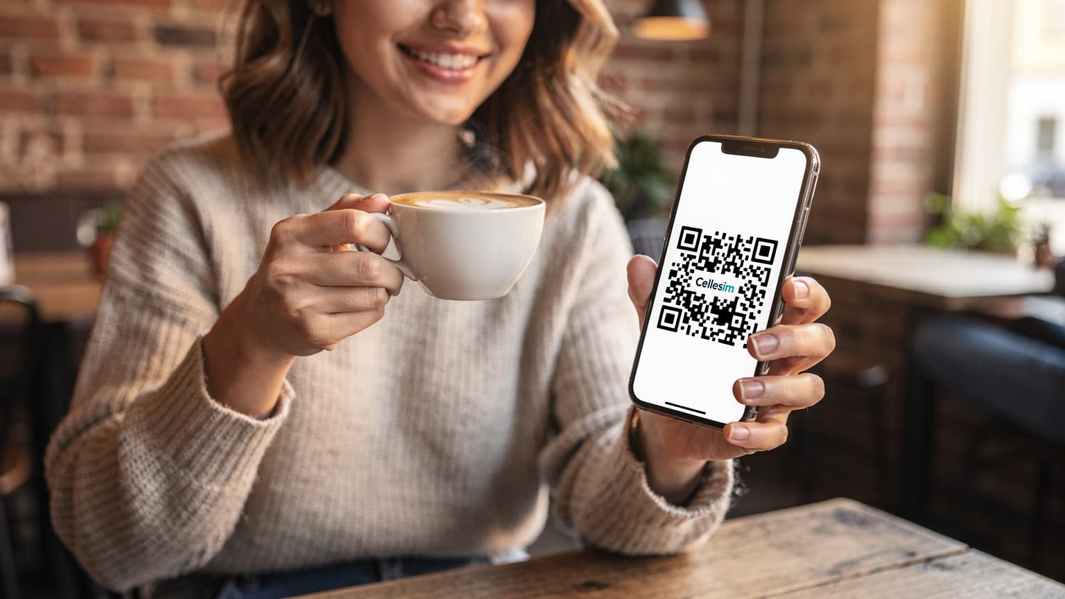 Eine junge Frau sitzt entspannt in einem Münchner Café im Glockenbachviertel, trinkt Cappuccino und hält ihr Smartphone in der Hand, auf dem sie einen Cellesim eSIM-QR-Code scannt.