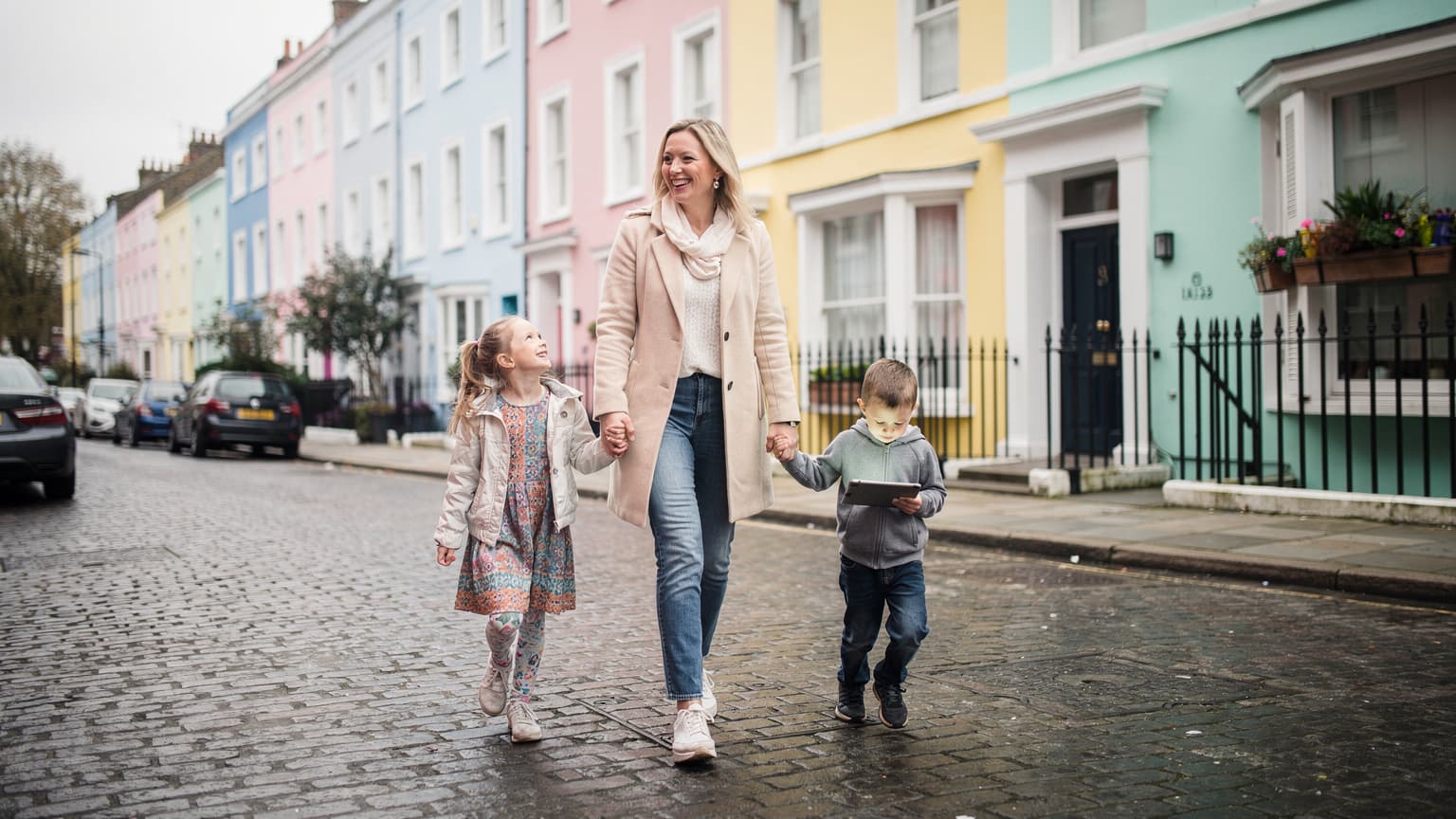 Una madre sonriente con sus dos hijos, uno de ellos con una tablet, caminando por una calle adoquinada en el barrio de Notting Hill, Londres, con casas de colores pastel al fondo.