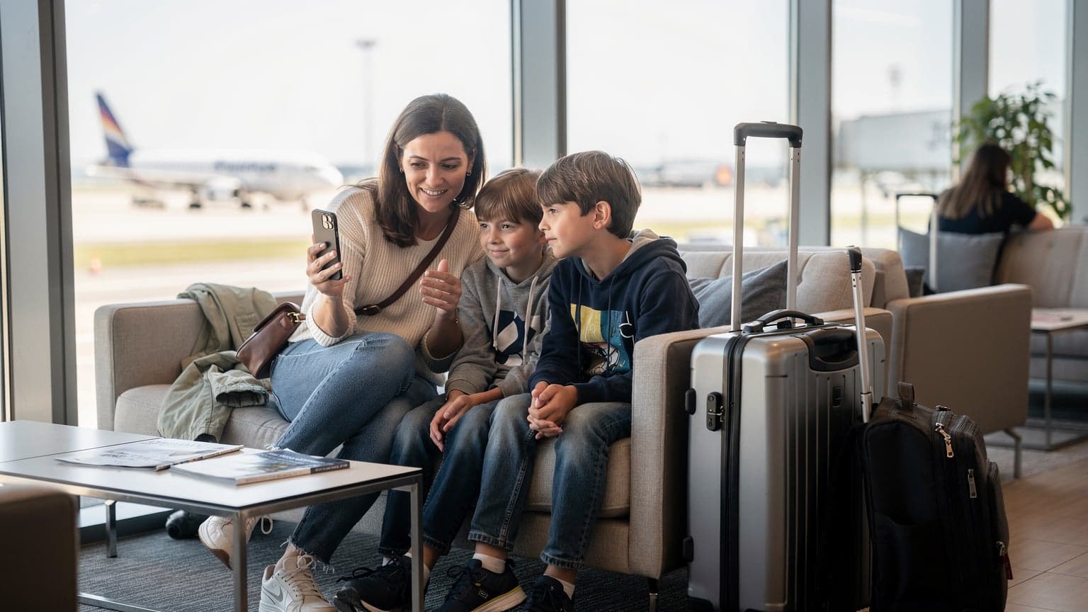 O mamă zâmbitoare, cu doi copii, privește ecranul unui iPhone nou, în timp ce se află într-un lounge modern de aeroport din Cluj-Napoca, cu bagaje de mână lângă ei.