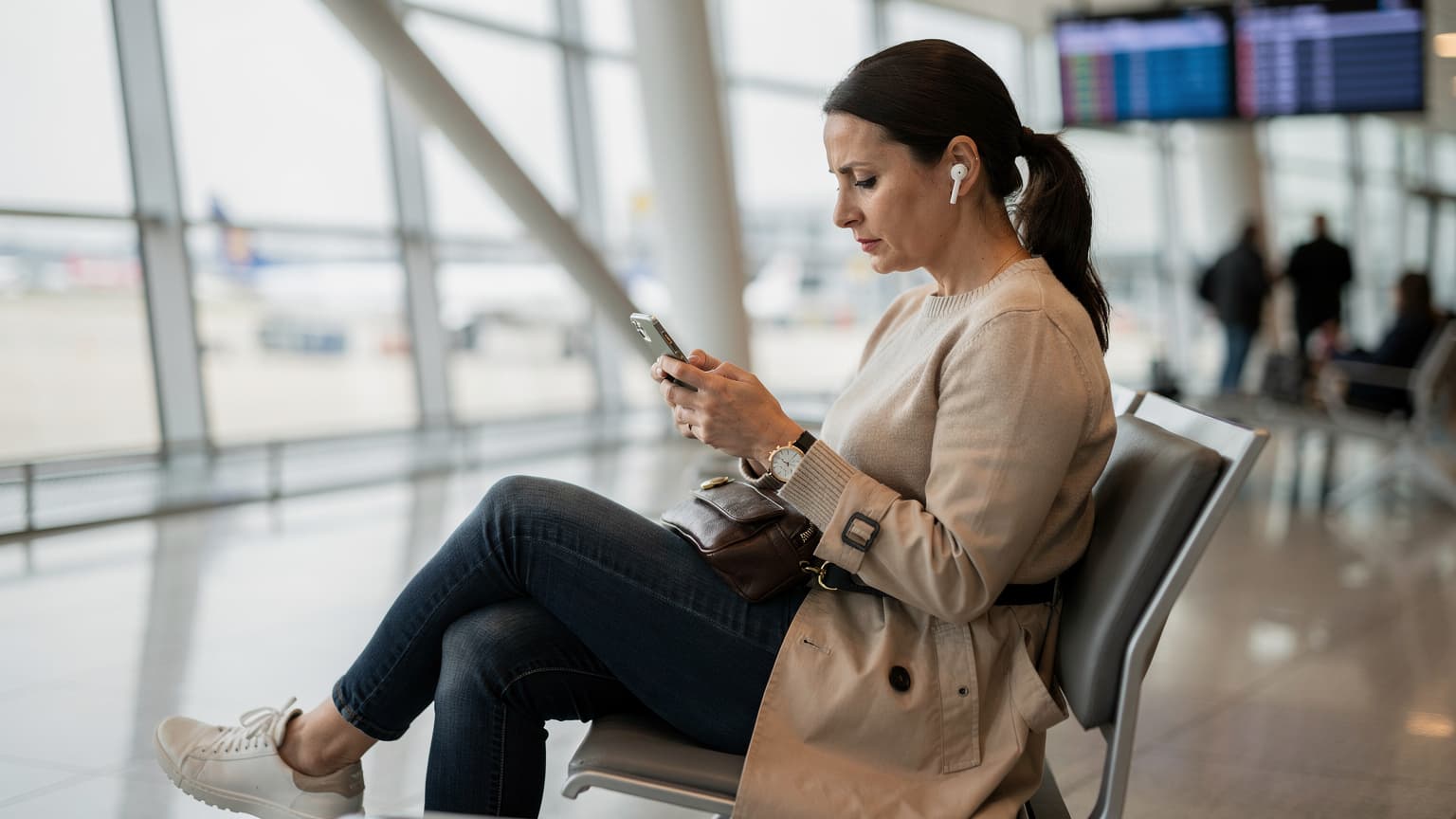 O femeie stă pe o bancă în Aeroportul Otopeni, București, verificând setările eSIM pe un iPhone, cu un pașaport deschis lângă ea și o geantă de laptop la picioare.