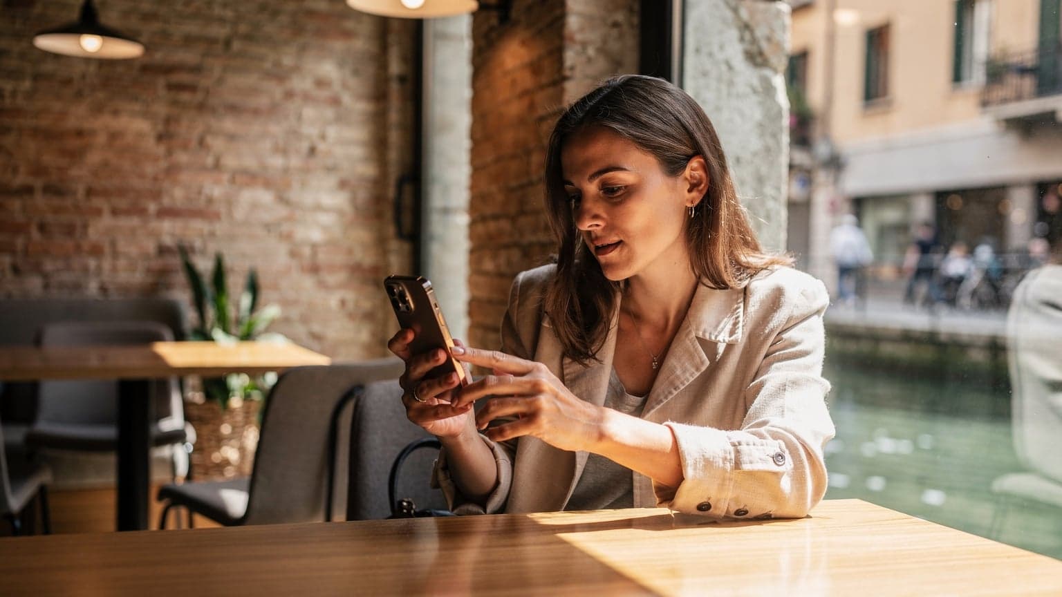Una donna seduta al tavolo di un caffè di Milano, con un iPhone in mano che mostra lo schermo, un passaporto e una tazza di caffè, mentre lavora o pianifica un viaggio.