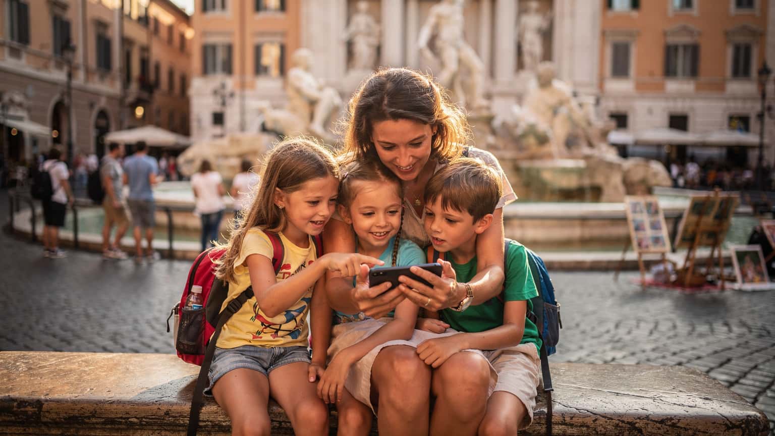 O mamă zâmbitoare, cu doi copii mici lângă ea, consultând un telefon cu ecranul luminat, pe o bancă într-o piață aglomerată din Roma, Italia, cu Fântâna Trevi în fundal.