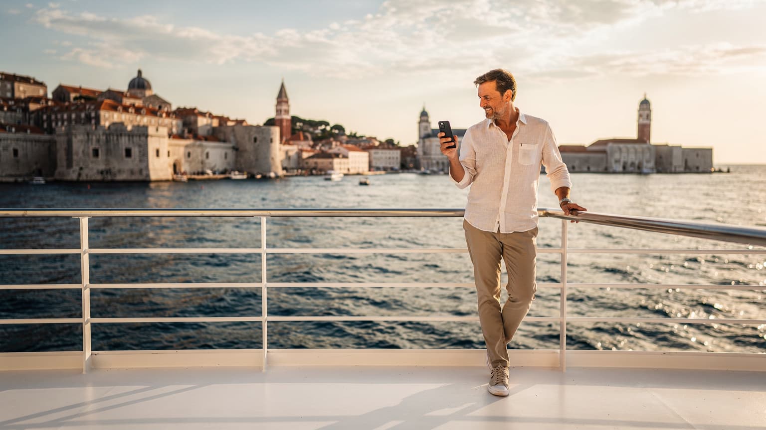 A person on the deck of a luxury cruise ship, holding a smartphone and looking out at the sunlit Adriatic Sea with a historic European coastal city in the distance, representing modern travel and seamless digital connectivity.