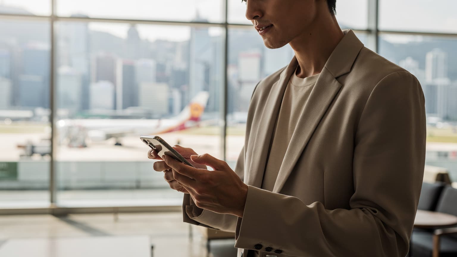 A person holding a smartphone in a modern, sunlit airport lounge in Hong Kong, with a city skyline or airplane visible through the window, conveying technology and travel.