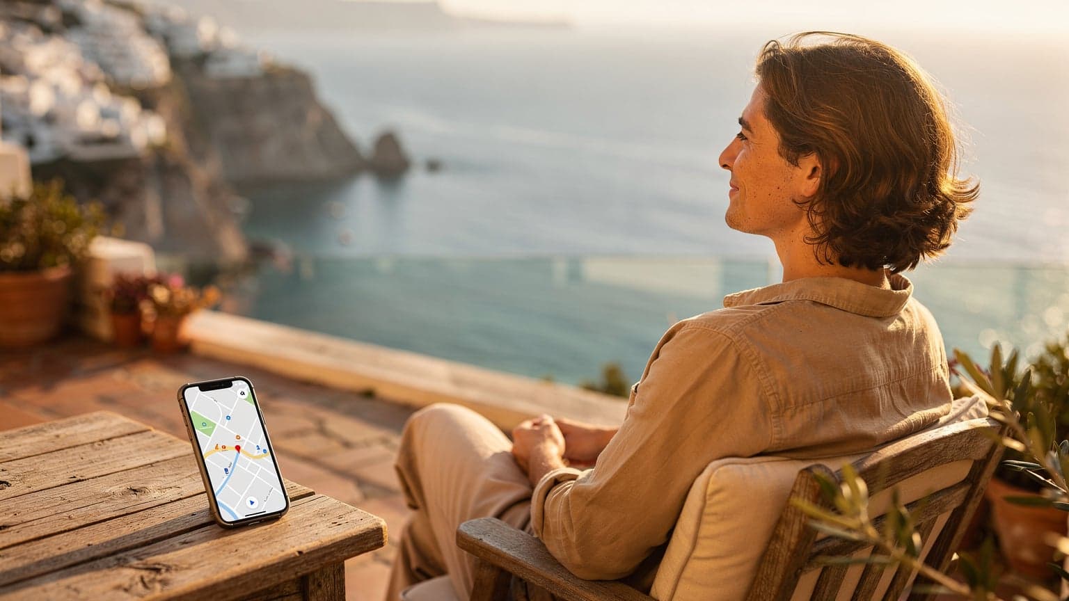 A person relaxing on a sunny terrace with a stunning view of the Mediterranean Sea at sunset. A generic smartphone displaying a map is subtly placed nearby, symbolizing carefree summer travel and seamless connectivity.