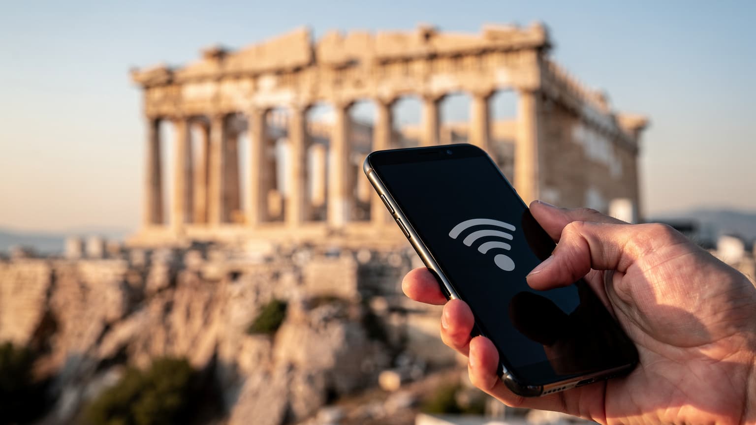 A hand holds a smartphone with a network signal, with the blurred Parthenon in Athens at golden hour in the background.