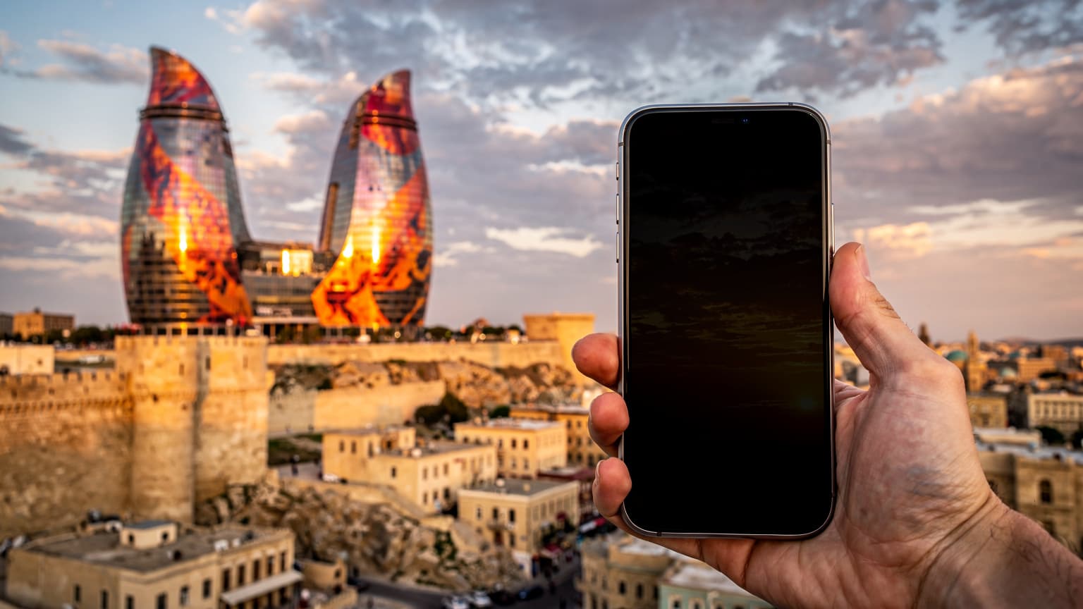A cinematic, photorealistic hero image for a travel and technology magazine. A modern smartphone is subtly held in the foreground, slightly out of focus. In the background, the iconic Flame Towers of Baku, Azerbaijan, rise against a vibrant golden hour sky, with historic Old City architecture visible below. The image conveys effortless, smart global connectivity while traveling.