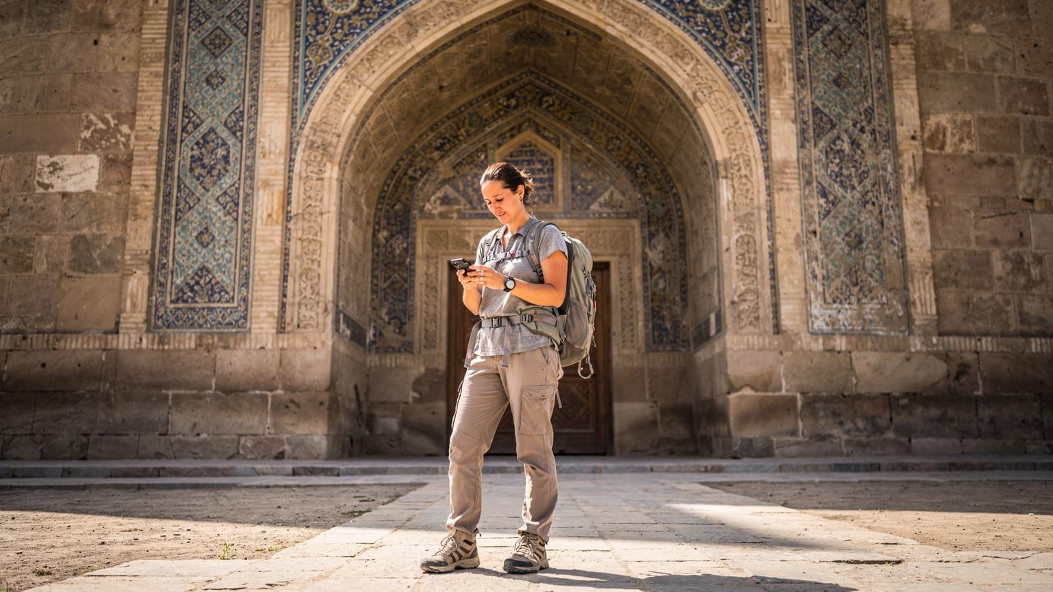 Una viajera, con mochila y sombrero, mira su teléfono mientras consulta un mapa digital frente a la entrada histórica de un caravanserai de piedra en Uzbekistán bajo un cielo azul claro.