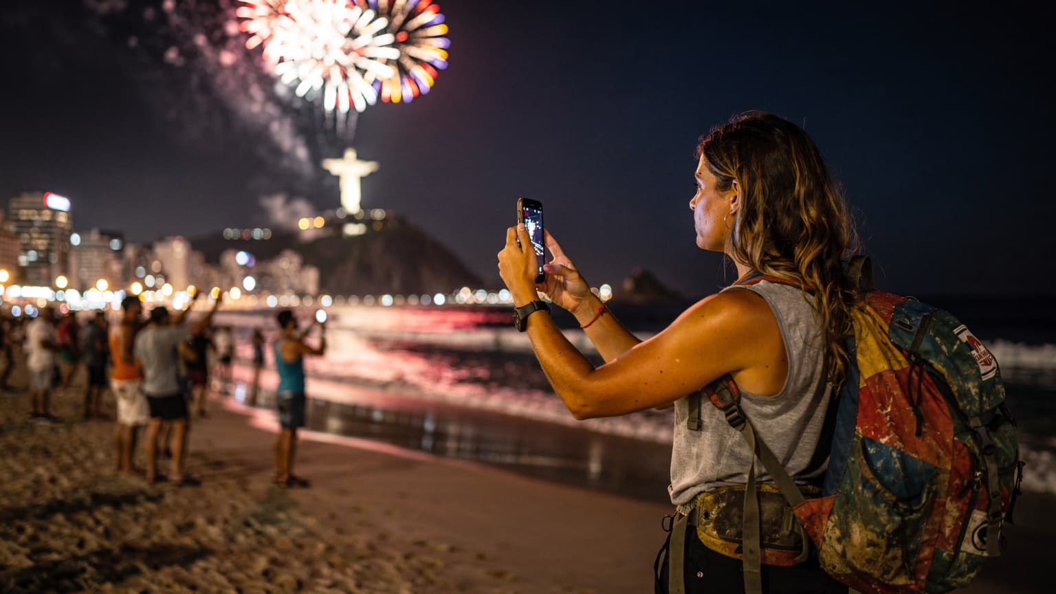 Uma mulher sorridente com uma mochila, usando o celular para tirar uma foto dos fogos de artifício da virada do ano em uma praia movimentada do Rio de Janeiro, com o Cristo Redentor ao fundo, iluminado.