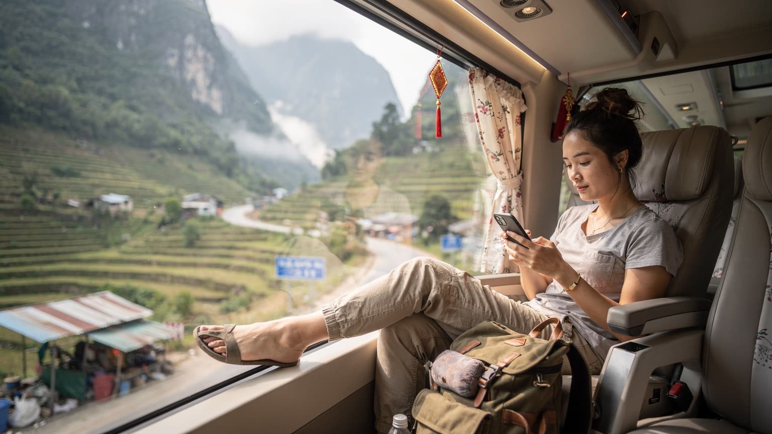 Một người phụ nữ trẻ đang kiểm tra điện thoại trên xe khách giường nằm cao cấp tại Việt Nam, khung cảnh núi non hùng vĩ lướt qua cửa sổ, ánh sáng ban mai dịu nhẹ chiếu vào.