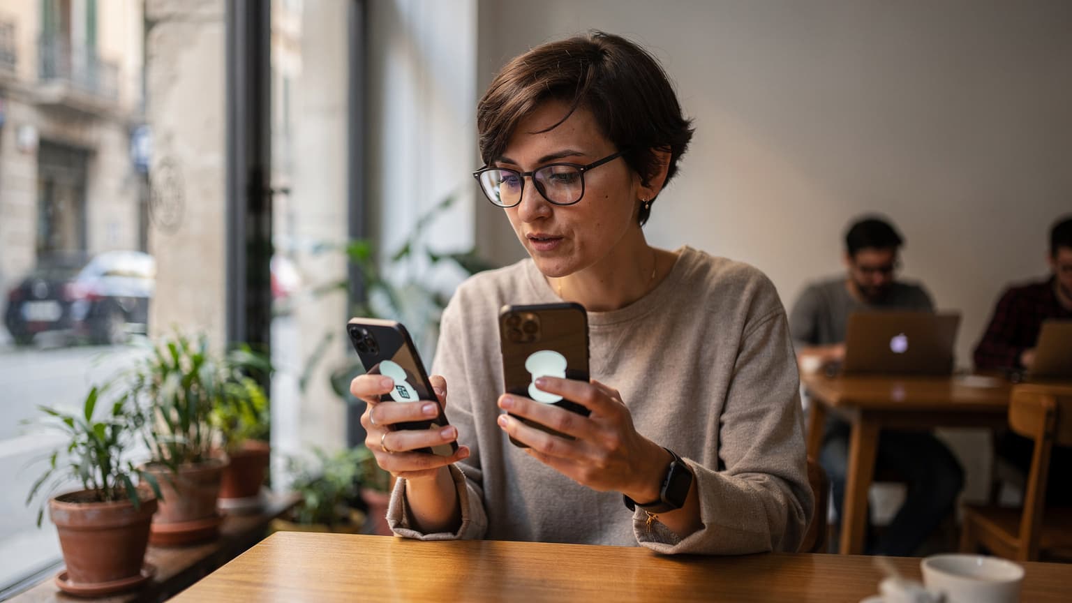 Une femme aux cheveux bruns, l'air concentré, tenant un nouvel iPhone et un ancien iPhone côte à côte, dans un café moderne et lumineux du quartier de Gràcia à Barcelone.