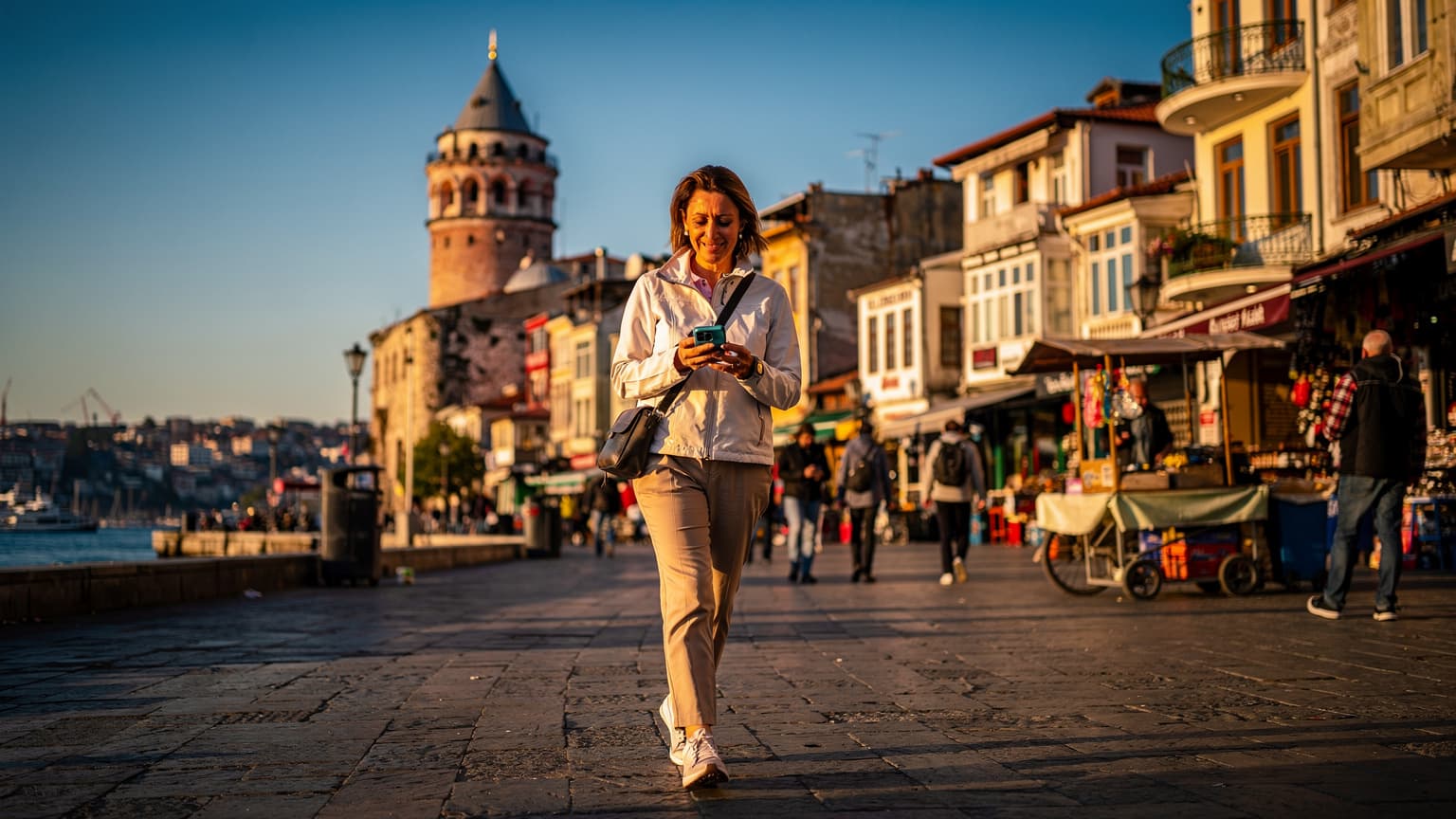 İstanbul'daki Karaköy iskelesinden Galata Kulesi'ne doğru yürüyen, elinde telefonuyla haritaya bakan bir gezgin, arka planda Boğaz ve tarihi binalar.
