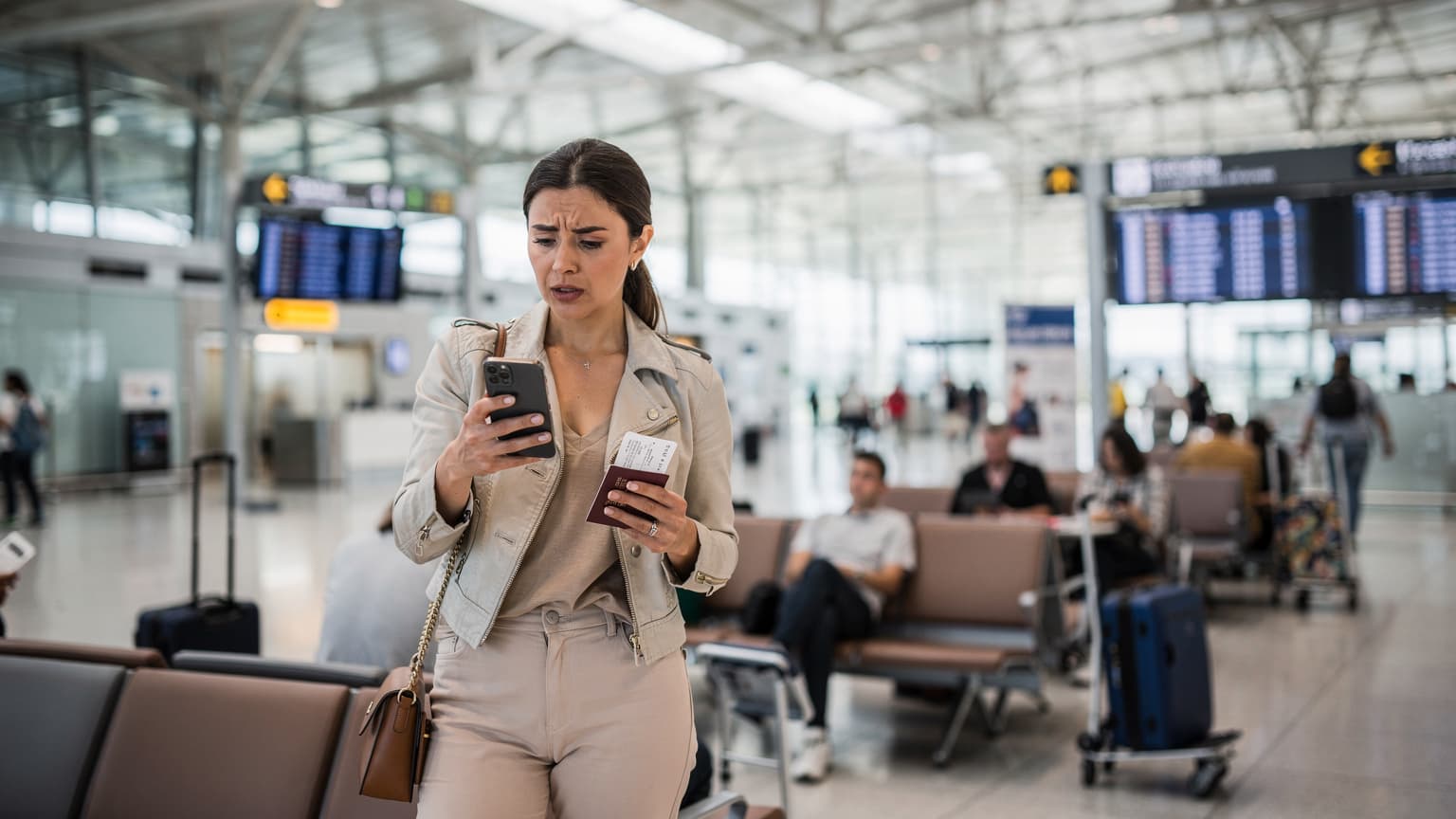 A woman at Charles de Gaulle Airport in Paris looks intently at her smartphone screen, holding a passport and a boarding pass, with the terminal's modern architecture in the background.