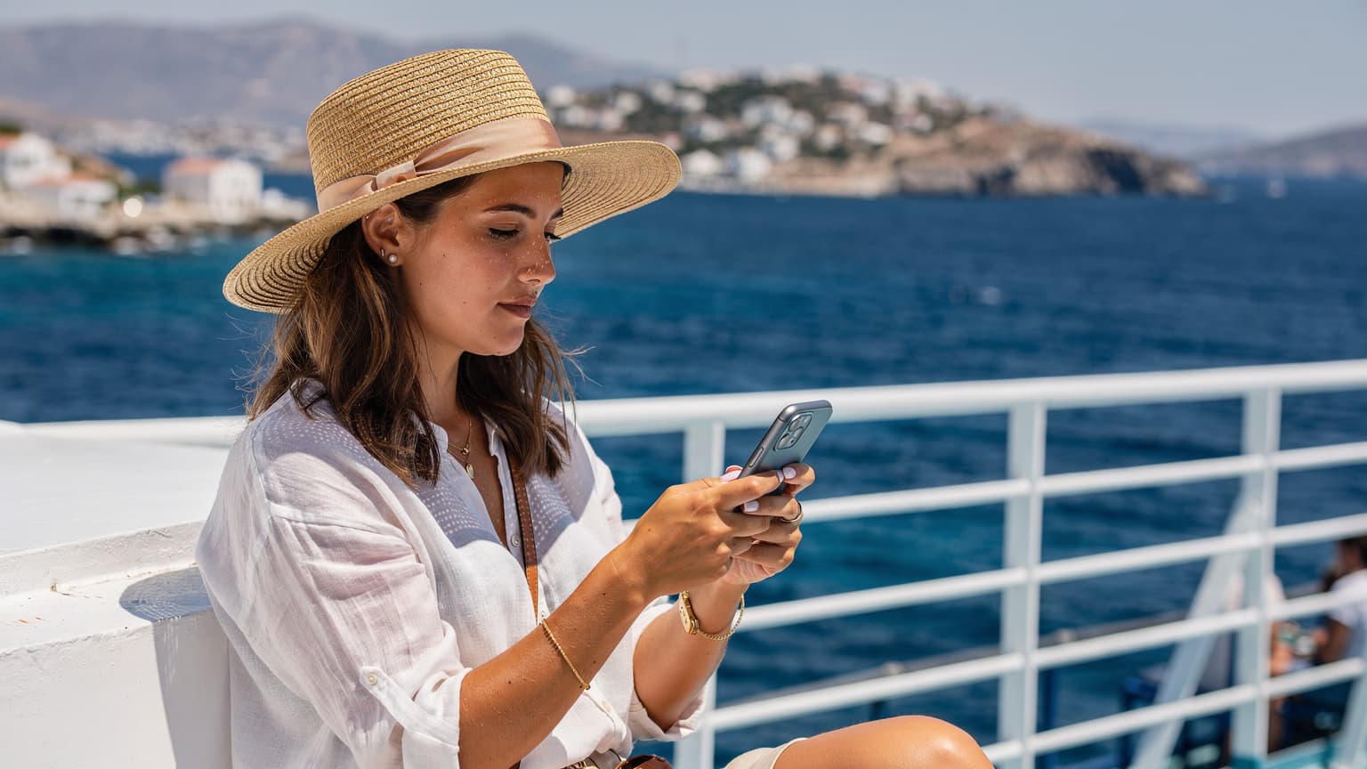En ung kvinde navigerer på sin smartphone på en solrig færge, mens den sejler gennem det azurblå Ægæiske Hav mellem græske øer. Hun smiler let, mens hun tjekker sin rute.