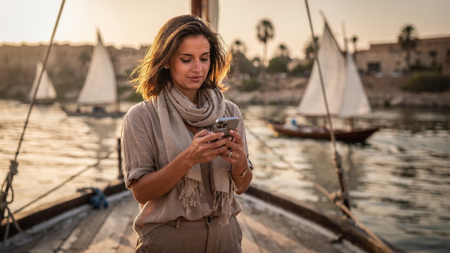 Une voyageuse naviguant sur son smartphone, le Nil et des felouques traditionnelles en arrière-plan à Louxor, sous un soleil couchant doré.