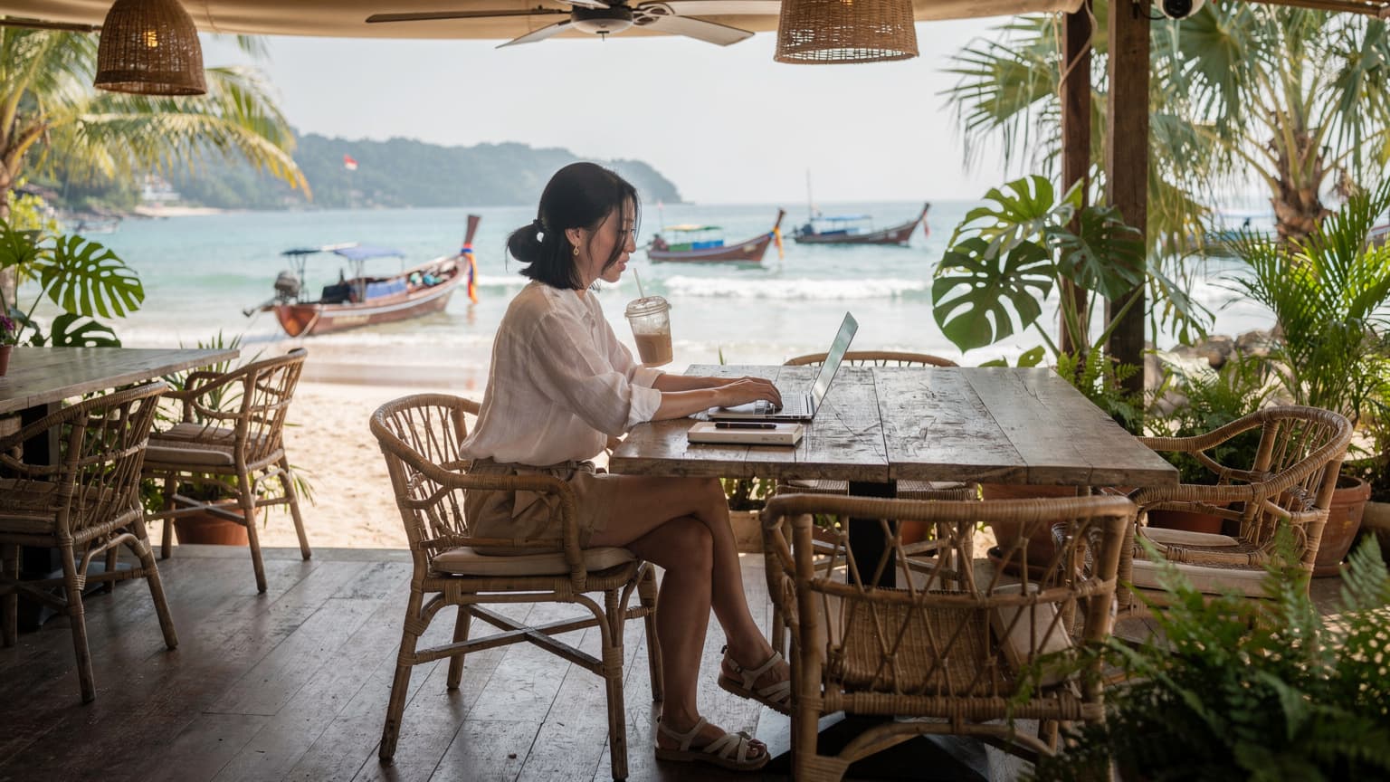 一名中国女性数字游民坐在普吉岛海滩边的咖啡馆里,面前放着笔记本电脑和一杯冰咖啡,背景是碧蓝的海水和棕榈树。